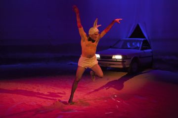 Pörröiseen kaniasuun pukeutunut henkilö tanssii energisesti punaisella valaistulla näyttämöllä, ja taustalla näkyy dramaattisessa valaistuksessa auto ja kaksi ihmistä sen sisällä.