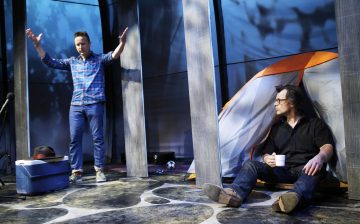 Lavalla on kaksi miestä, joista toinen seisoo ja elehtii kädet ylhäällä kylmälaukun vieressä, kun taas toinen istuu teltan vieressä valkoinen muki kädessään ja katsoo seisovaa miestä kohti. Kohtauksessa on retkeilyteema ja dramaattinen valaistus.