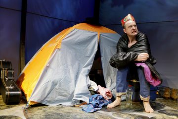 Mies, jolla on tilapäinen kruunu ja makuupussi, istuu oranssin teltan ulkopuolella vaatteiden ja vesipullon ympäröimänä, ja lähellä on kitaralaukku ja saappaat. Hän näyttää tekevän dramaattista ilmettä.