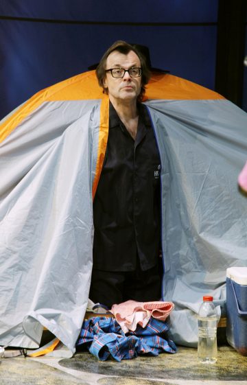 Silmälasipäinen ja mustapaitainen mies seisoo osittain oranssin ja harmaan teltan sisällä. Hänen edessään on erilaisia esineitä, kuten ruutupaita, kangas ja muovipullo.