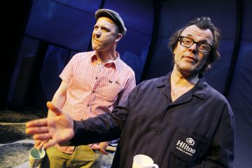 Kaksi miestä seisoo sisätiloissa mukeja kädessään. Oikeanpuoleinen mies, jolla on silmälasit ja tumma takki, elehtii kädellään puhuessaan. Vasemmalla olevalla miehellä on vaalea paita ja lippis, ja hän katsoo mietteliäästi kaukaisuuteen.