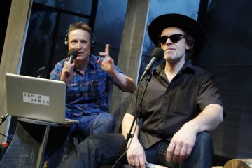 Kaksi miestä istuu mikrofonien edessä; toisella on kuulokkeet ja hän elehtii puhuessaan, toisella on aurinkolasit ja leveälasinen hattu. Heidän edessään pöydällä on kannettava tietokone, jossa lukee RADIO OKNAS.