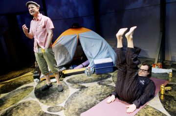 Kaksi miestä lavalla: toinen seisoo ja puhuu rennosti pukeutuneena teltan ja retkeilyvarusteiden äärellä; toinen makaa matolla jalat koholla, silmälasit päässä ja tummissa vaatteissa, ja näyttää harjoittelevan tai venyttelevän.