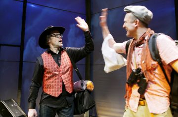 Kaksi miestä antaa lavalla toisilleen high-fivea. Toisella on musta hattu, silmälasit ja punainen liivi, kun taas toisella on harmaa baretti, oranssi huivi ja ruutupaita, ja hänellä on kamera ja muovipussi kädessään.