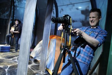 Ruudulliseen paitaan pukeutunut mies säätää jalustalla olevaa kameraa ja tekee jännittyneen ilmeen. Taustalla toinen hattupäinen mies soittaa kitaraa oranssin teltan lähellä sinisellä valaistulla näyttämöllä.