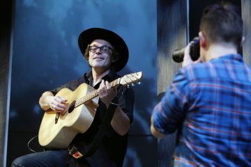 Silmälasipäinen ja leveälasipäinen hattuinen henkilö soittaa akustista kitaraa ja hymyilee, kun toinen ruutupaitainen henkilö kuvaa heitä studiossa.