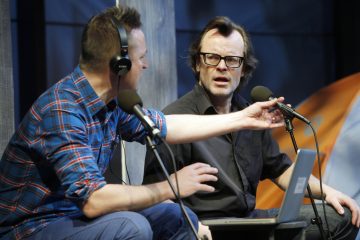 Kaksi miestä istuu pöydän ääressä mikrofonien ja kuulokkeiden kanssa keskustellen. Toinen ruudulliseen paitaan pukeutunut mies elehtii, kun taas toinen, silmälasipäinen ja mustiin pukeutunut mies näyttää yllättyneeltä. Kannettava tietokone on avoinna heidän edessään.