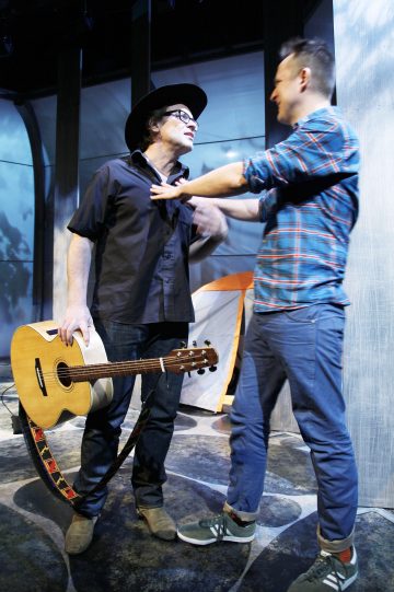 Lavalla on kaksi miestä vuorovaikutuksessa; toinen mustaan paitaan ja cowboy-hattuun pukeutuneista pitelee akustista kitaraa, kun taas toinen sinikuvioiseen ruutupaitaan pukeutunut mies työntää varovasti rintaansa. Molemmat näyttävät keskustelevan.