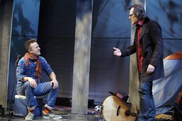 Kaksi miestä on näyttämöllä dramaattisessa kohtauksessa. Toinen istuu kylmälaukussa farkut ja ruutupaita yllään, kun taas toinen seisoo ja elehtii kädellään. Lähistöllä näkyy kitara, hattu ja oranssivalkoinen teltta.