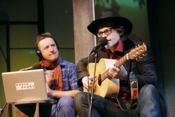 Kaksi miestä esiintyy lavalla. Toinen laulaa ja soittaa akustista kitaraa ja käyttää mustaa hattua ja silmälaseja. Toinen, huiviin ja ruutupaitaan pukeutunut mies istuu kannettavan tietokoneen vieressä, jossa lukee RADIO OKNAŚ, ja katselee ja kuuntelee.