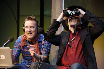 Kaksi miestä istuu vierekkäin mikrofonien edessä; toisella on kuulokkeet ja hän elehtii puhuessaan, toisella on hattu ja hän käyttää kiikaria. Heidän edessään pöydällä on kannettava tietokone.