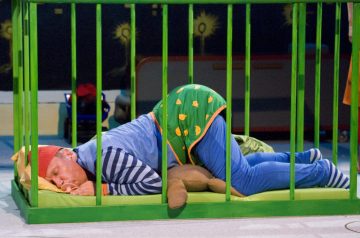 Värikkääseen, leikkisään pyjamaan ja punaiseen yömyssyyn pukeutunut henkilö nukkuu käpertyneenä vihreässä pinnasängyssä, halaten pehmokarhua keltaisella tyynyllä, omituisessa, lapsenomaisessa ympäristössä.