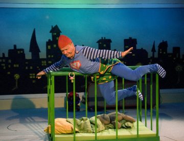 Värikkääseen pukuun pukeutunut esiintyjä hyppää lavalla kirkkaanvihreän pinnasängyn yli, jonka sisällä on nukke ja huopa ja takana kaupunkimaisema.
