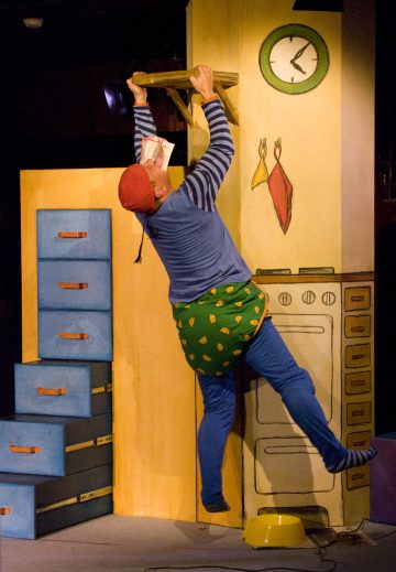 Värikkäisiin, leikkisiin vaatteisiin pukeutunut henkilö roikkuu puuhyllyllä, joka on kiinnitetty keittiötä muistuttavaan maalattuun lavasteeseen, jonka taustalla näkyvät kello, uuni ja kaapit.