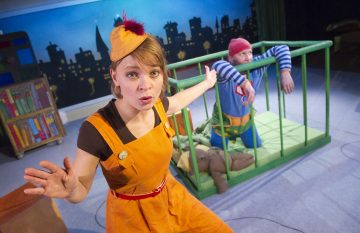 Oranssiin asuun ja hattuun pukeutunut nainen seisoo etualalla kädet ojennettuina, kun taas värikkäisiin vaatteisiin ja punaiseen lippikseen pukeutunut mies istuu vihreässä sängyssä taustalla kaupunkimaisemaa vasten.