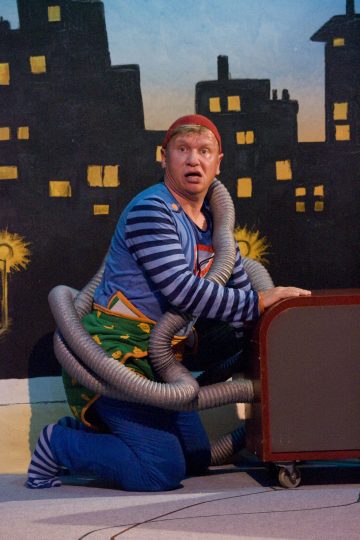 Värikkääseen pukuun pukeutunut mies polvistuu lavalla yllättynyt ilme kasvoillaan, käärittynä joustaviin harmaisiin putkiin, ja taustalla on maalattu kaupunkikuva, jossa on keltaisesti valaistuja ikkunoita.