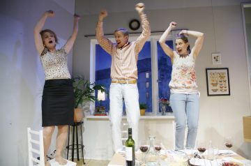 Kolme ihmistä seisoo ruokapöydän ympärillä olevilla tuoleilla, nostavat kätensä ylös ja hurraavat. He vaikuttavat iloisilta ja innostuneilta. Pöydällä on viinilaseja ja ruokaa, ja taustalla on ikkuna ja kasveja.