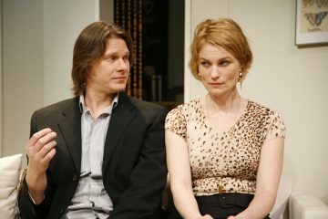 Mustaan bleiseriin pukeutunut mies ja leopardikuvioiseen toppiin pukeutunut nainen istuvat vierekkäin sohvalla, ja molemmat katsovat sivulle epäilevästi tai hämmentyneinä.