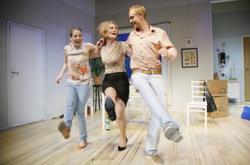 Kolme ihmistä tanssii iloisesti kirkkaasti valaistussa huoneessa, jossa on puulattia. He yhdistävät kätensä ja potkivat jalkojaan yhtä aikaa hymyillen ja nauraen. Tilassa on hajallaan huonekaluja, laatikoita ja erilaisia kotitaloustavaroita.