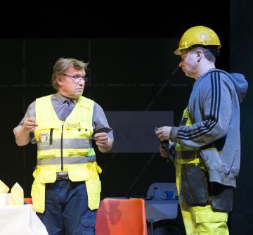 Kaksi keltaisiin turvaliiveihin ja suojakypärään pukeutunutta miestä keskustelee sisätiloissa. Toisella miehellä on kädessään pieni esine; he seisovat lähellä pöytää, jossa on papereita ja raidallisia esineitä tummalla taustalla.