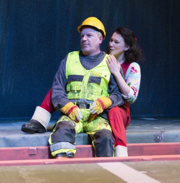 Keltaiseen työasuun ja kypärähattuun pukeutunut mies istuu jalkakäytävällä, ja punaisiin haalareihin ja kukkapaitaan pukeutunut nainen istuu hänen vieressään ja laskee kätensä miehen olkapäälle. Molemmat näyttävät olevan syvällä ajatuksissaan.