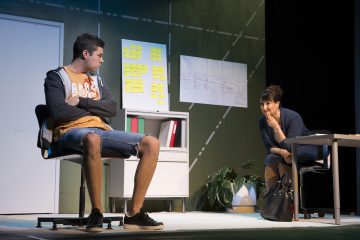 Nuori mies istuu rennosti pukeutuneena, kädet ristissä, vastapäätä vanhempaa naista työpöydän ääressä toimistoympäristössä, jossa on muistilappuja, taulukoita seinällä ja arkistokaappi lähellä. Nainen näyttää kuuntelevan mietteliäästi.