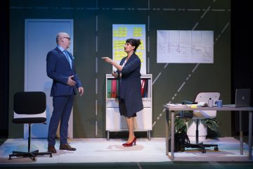 Kaksi ihmistä seisoo toimistoympäristössä lavalla. Siniseen pukuun pukeutunut mies kuuntelee, kun laivastonsiniseen mekkoon ja punaisiin korkokenkiin pukeutunut nainen elehtii. Taustalla on muistilappuja, papereita seinällä ja toimistokalusteita.