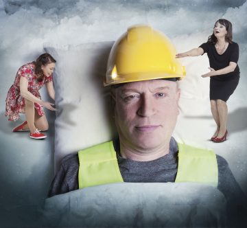 Mies, jolla on keltainen suojakypärä ja turvaliivi, makaa tyynyllä, ja kaksi pienoiskoossa olevaa naista, toinen kukkamekkoon ja toinen mustaan pukeutuneena, näyttävät seisovan hänen päänsä molemmin puolin ja elehtivän ilmeikkäästi.