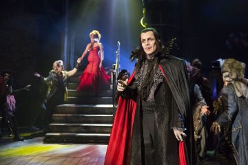 Lavalla seisoo dramaattiseen mustapunaiseen vampyyriasuun pukeutunut mies, jota ympäröivät hienostuneisiin asuihin pukeutuneet näyttelijät. Punapukuinen nainen seisoo miehen takana portailla teatterivalaistuksessa.