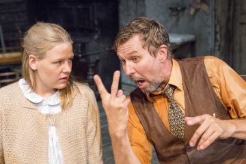 Mies, jolla on ruskea liivi ja oranssi paita, elehtii vilkkaasti kahdella sormella ja puhuu vaaleahiuksiselle naiselle, jolla on beige pusero ja joka katsoo häntä hämmentyneenä.