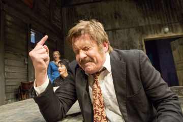 Pukuun ja kuviolliseen solmioon pukeutunut mies istuu pöydän ääressä, silmät tiukasti kiinni ja kasvot jännittyneinä, ja nostaa kätensä sormet puristettuina. Taustalla on kolme ihmistä, jotka vaikuttavat yllättyneiltä tai huolestuneilta hämärästi valaistussa lavastuksessa.