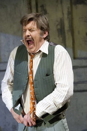 Vihreään liiviin, raidalliseen paitaan ja oranssikuvioiseen solmioon pukeutunut mies seisoo sisätiloissa silmät kiinni ja suu auki, ja hän näyttää huutavan tai reagoivan tuskissaan puristaessaan housujaan vyötärönsä lähellä.