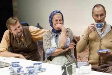 Kolme aikuista istuu pöydän ääressä, jossa on sinivalkoisia teekuppeja. Keskellä istuva nainen, jolla on päähuivi, hymyilee käsi suunsa edessä. Molemmin puolin istuvat miehet vaikuttavat rentoutuneilta; yksi juo teekupista.