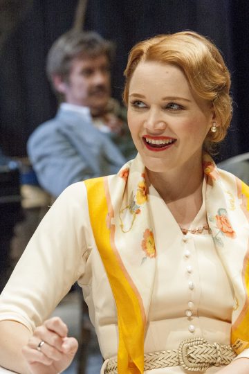 Nainen, jolla on punaiset, vintage-aaltoiksi muotoillut hiukset, valkoinen mekko ja kukkahuivi, jossa on keltainen reunus, hymyilee istuessaan sisätiloissa. Taustalla on hämärtynyt pukuun pukeutunut mies.