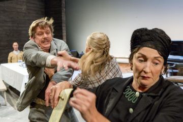 Mies kurottautuu aggressiivisesti naista kohti, kun taas toinen nainen etualalla katsoo huolestuneena poispäin. Taustalla istuva mies tarkkailee pöydän ääressä hämärässä, dramaattisessa ympäristössä.