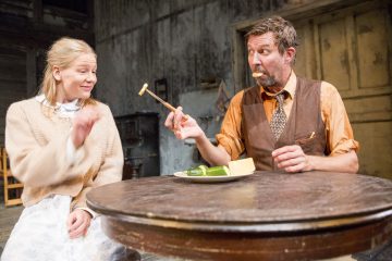 Mies ja nainen istuvat puupöydän ääressä. Mies pitelee haarukalla ruokapalaa ja tekee hassun naaman, kun taas nainen hymyilee. Heidän välissään lautasella on voita ja kurkkua. Asetelma näyttää maalaismaiselta ja vanhanaikaiselta.