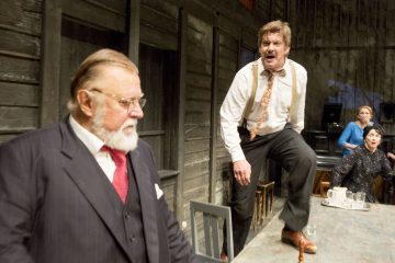 Vanhempi pukumies seisoo puupöydän vieressä, kun taas paitainen ja solmioinen mies huutaa vihaisena pöydän päällä. Taustalla istuu kaksi naista, jotka katselevat dramaattista kohtausta.