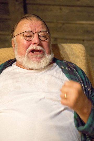 Sisällä istuu vanhempi mies, jolla on silmälasit, valkoinen parta ja ruudullinen paita valkoisen t-paidan päällä, ja hän puhuu kiihkeästi käsi eleenä kohotettuna.