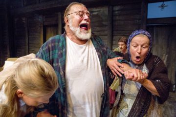 Valkoiseen paitaan ja ruudulliseen kaapuun pukeutunut vanhempi mies huutaa, kun huiviin pukeutunut nainen pitelee hänen kättään ja vaikuttaa järkyttyneeltä. Tyttö kumartaa päätään lähistöllä; taustalla seisoo kaksi muuta ihmistä maalaismaisessa puuhuoneessa.