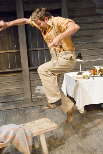 Ruskehtaviin housuihin ja oranssiin paitaan pukeutunut mies hyppää ilmaan lasipullo kädessään puisen penkin vieressä. Hänen takanaan on pöytä, jossa on lamppu, kirjoja ja pulloja ja joka on asetettu maalaismaista puuseinää vasten.