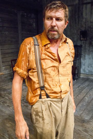 Mies, jolla on märät hiukset ja läpimärät vaatteet, oranssi paita, jossa on henkselit, ja beiget housut, seisoo sisällä puulattialla ja näyttää huolestuneelta.