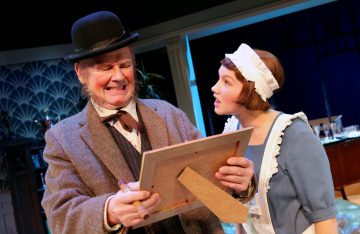 Vanhempi mies vintage-vaatteissa ja keilahattu päässä irvistää pitelemässä kuvakehystä, kun nuori nainen palvelijan univormussa katsoo häntä puhuen kirkkaasti valaistussa lavastuksessa.