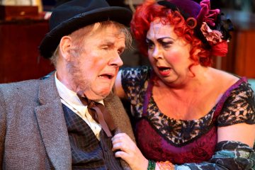 Vanhempi mies, jolla on hattu ja puku, näyttää ahdistuneelta, kun violettiin pitsimekkoon pukeutunut nainen, jolla on kiharat punaiset hiukset ja dramaattinen meikki, pitelee hänen olkapäätään ja katsoo häntä huolestuneena.