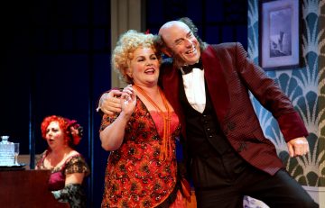 Kaksi värikkäisiin vintage-asuihin pukeutunutta näyttelijää hymyilee ja syleilee lavalla, kun punatukkainen nainen soittaa pianoa taustalla. Lavastuksessa on kuviollisia tapetteja ja kehystettyjä kuvia, jotka viittaavat teatteriesitykseen.