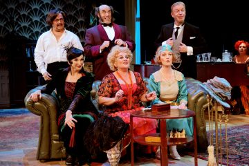 Kuusi värikkäisiin aikakauden pukuihin pukeutunutta näyttelijää istuu ja seisoo vihreän sohvan ympärillä teatterilavalla, vaikuttavat sitoutuneilta ja ilmeikkäiltä, ja taustalla näkyy rekvisiittaa ja vintage-koristeita.