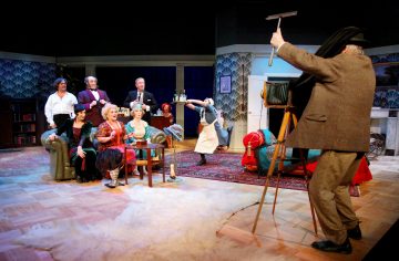 Ryhmä vintage-vaatteisiin pukeutuneita ihmisiä poseeraa koristellulla lavasteella, kun valokuvaaja vanhanaikaisella kameralla valmistautuu ottamaan heistä kuvan. Yksi henkilö ottaa dramaattisen poseerauksen, mikä lisää huumoria kohtaukseen.