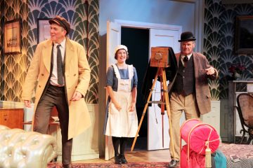 Lavalla esiintyy kolme näyttelijää vintage-asuissa; piian asuun pukeutunut nainen seisoo kahden pukuihin ja hattuihin pukeutuneen miehen välissä. Toinen mies pitää jalustalla olevaa kameraa. Lavastus muistuttaa eleganttia, vanhanaikaista olohuonetta.