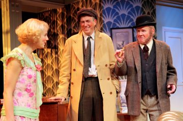 Lavalla on kolme vintage-asuihin pukeutunutta näyttelijää: kukkamekkoon pukeutunut nainen ja kaksi takkiin ja hattuihin pukeutunutta miestä seisovat yhdessä hymyillen ja keskustellen art deco -tyylistä taustaa vasten, jossa on kuviollinen tapetti.