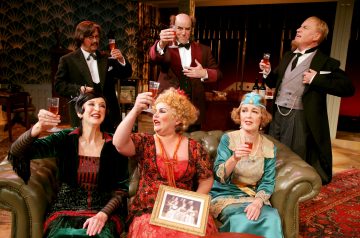 Kuusi tyylikkäisiin vintage-asuihin pukeutunutta aikuista istuu ja seisoo sohvan ympärillä ja kohottaa lasit maljan kohotukseksi. Edessä istuu kolme naista, joista toisella on kehystetty valokuva kädessään, ja heidän takanaan seisoo kolme virka-asuihin pukeutunutta miestä.