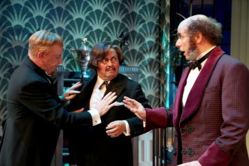 Kolme muodollisiin pukuihin pukeutunutta miestä käy dramaattista keskustelua. Yksi mies viittoo kädellään toisen rinnalle, kun taas kolmas näyttää yllättyneeltä. He seisovat tyylikkäässä huoneessa, jossa on kuviollinen tapetti.